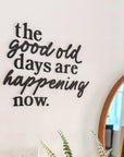 Decorative wall quote 'the good old days are happening now.' with a plant on a table.