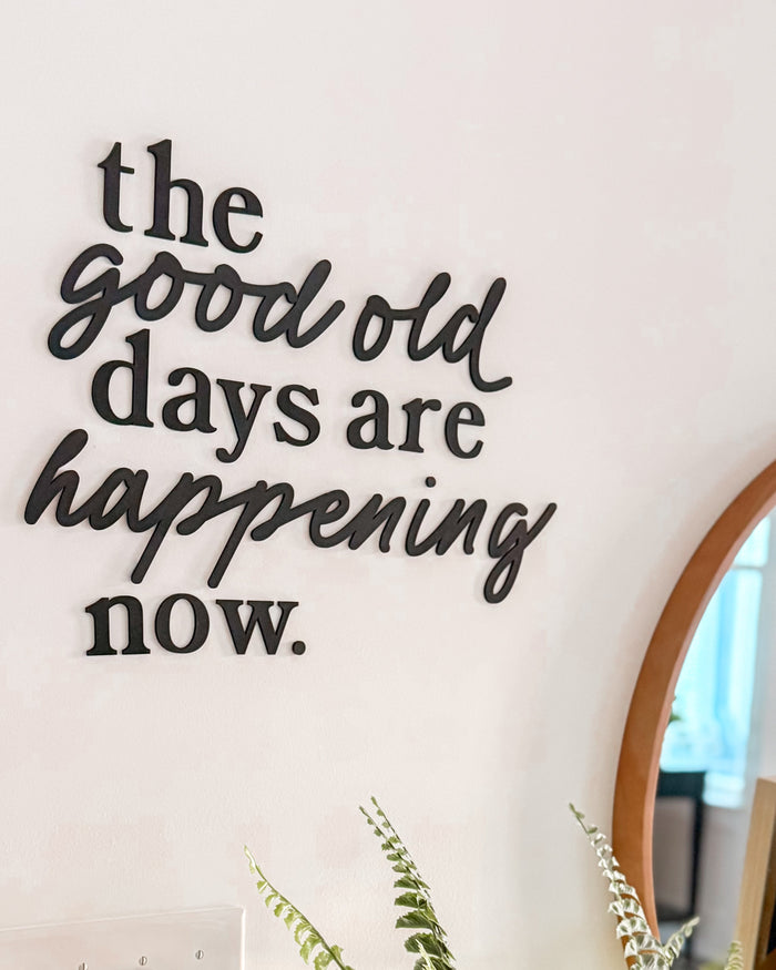 Decorative wall quote 'the good old days are happening now.' with a plant on a table.