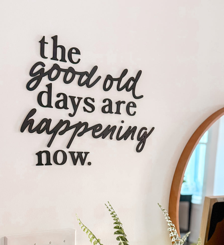 Decorative wall quote 'the good old days are happening now.' with a plant on a table.