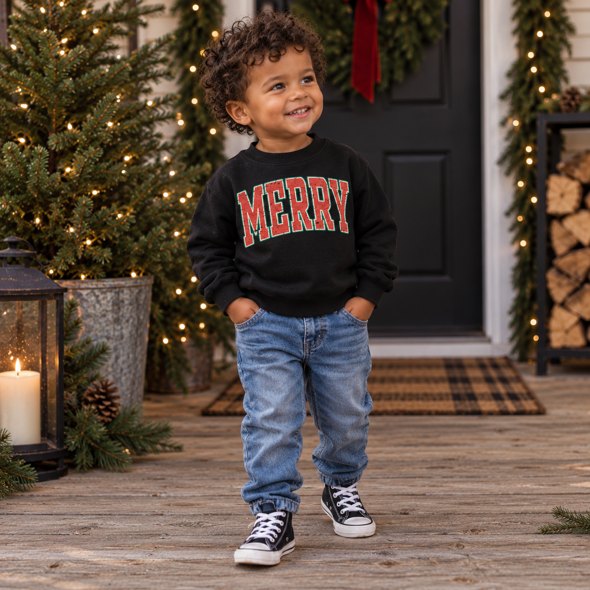 A toddler in a black MERRY sweatshirt and jeans on a front porch decorated for Christmas